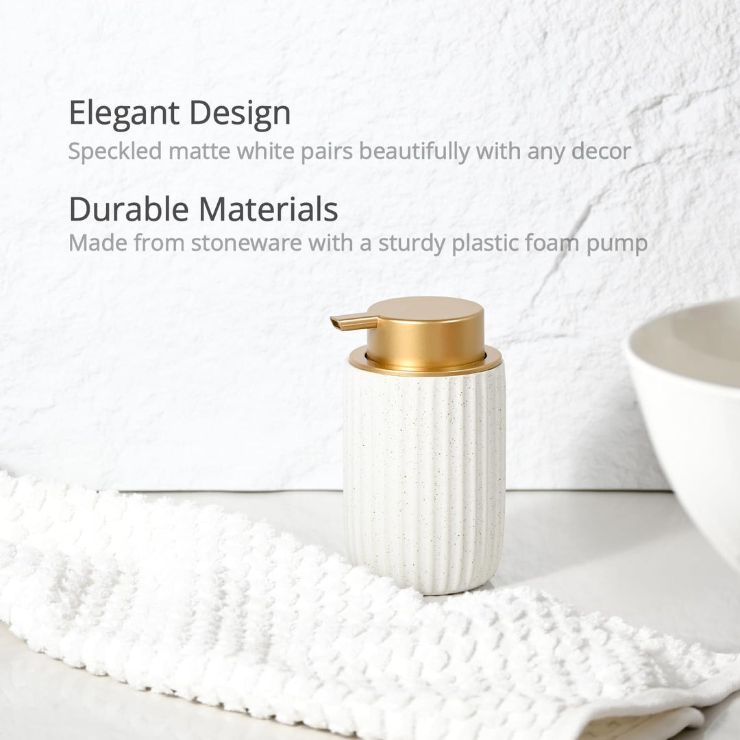 Speckled matte white stoneware dispenser with a gold foam pump on a white counter beside a woven towel