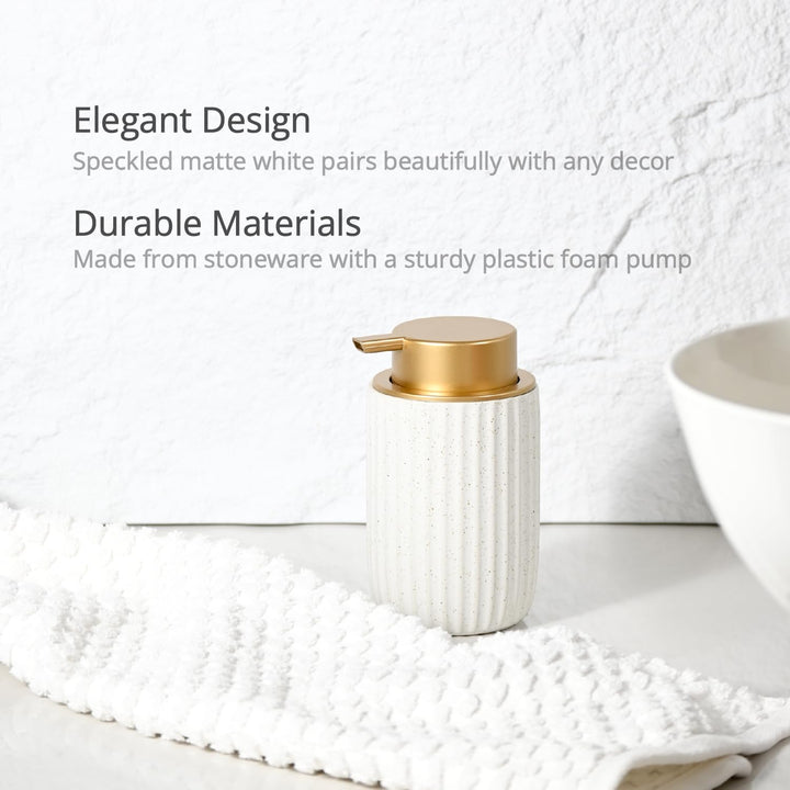 Speckled matte white stoneware dispenser with a gold foam pump on a white counter beside a woven towel