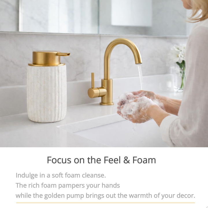 Speckled white ceramic soap dispenser beside a gold faucet as hands are washed with foam at a white sink